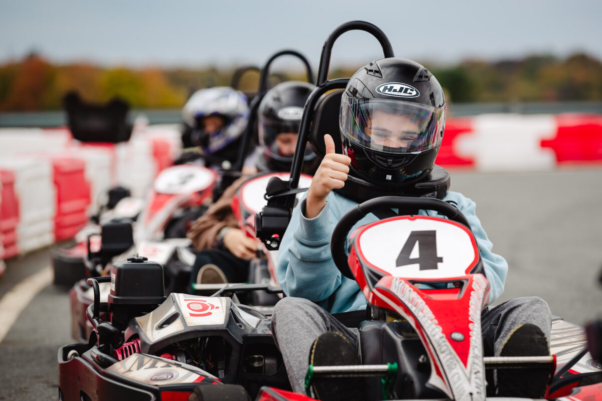 A group of teenagers are sitting in go karts with helmets on. The first teen is giving the thumbs up