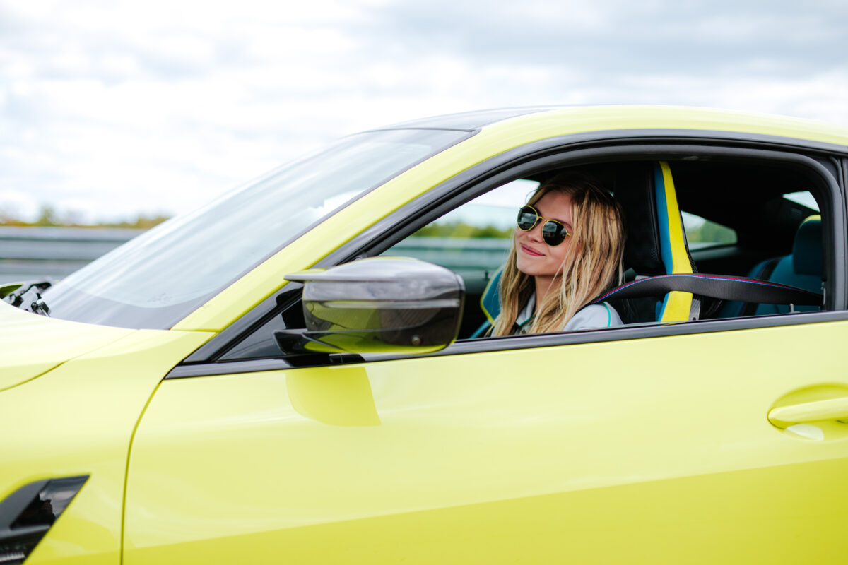 A blonde female sitting in a green/yellow BMW M3 Competition, smiling at the camera