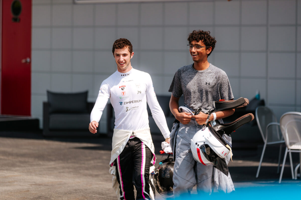 Two young men in racing suits are walking through pit lane holding their helmets