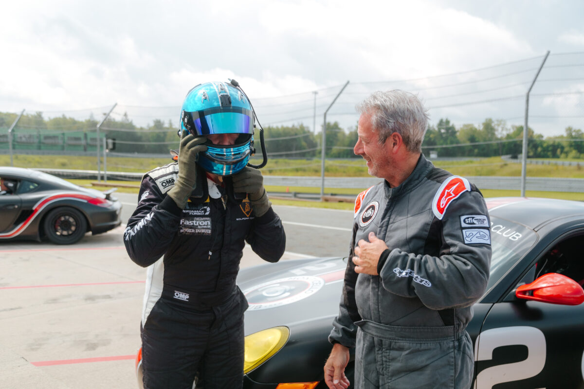 Two drivers in racing suits are standing in front of a race car in pit lane