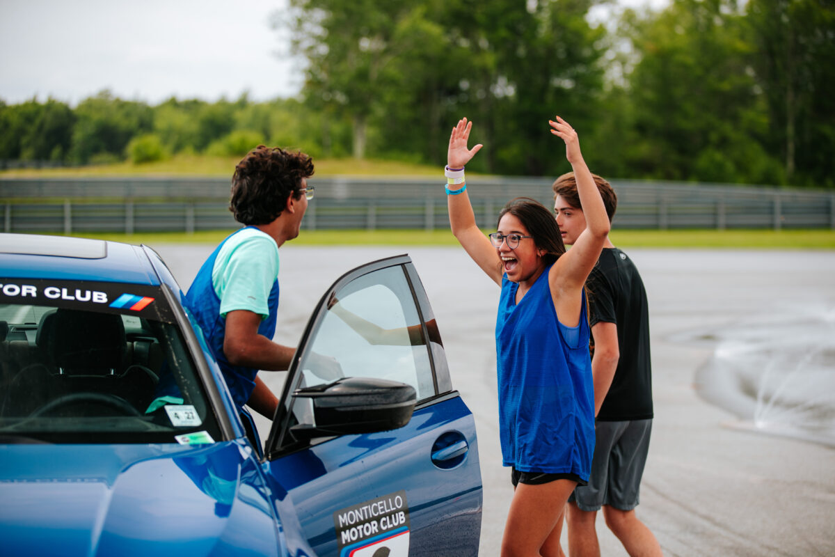 A female teen is standing next to a car with her hands up in the air and an excited look on her face