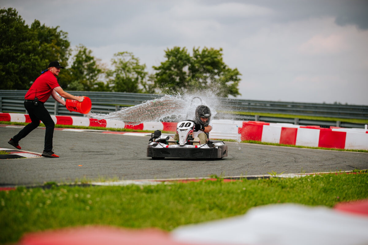 A teenager is racing in a go kart as an instructor is pouring a big bucket of water at them