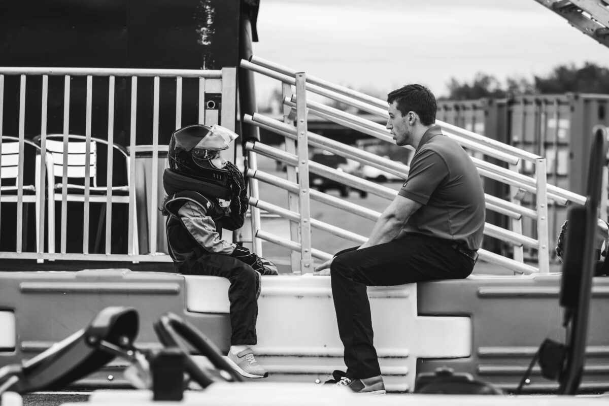A black & white image of an instructor speaking to a child in a race suit and helmet at the Karting Facility