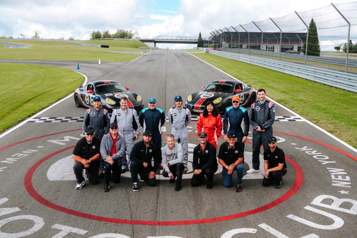 A group photo of race school students and instructors, posing on the start/finish line with two Porsches in the background