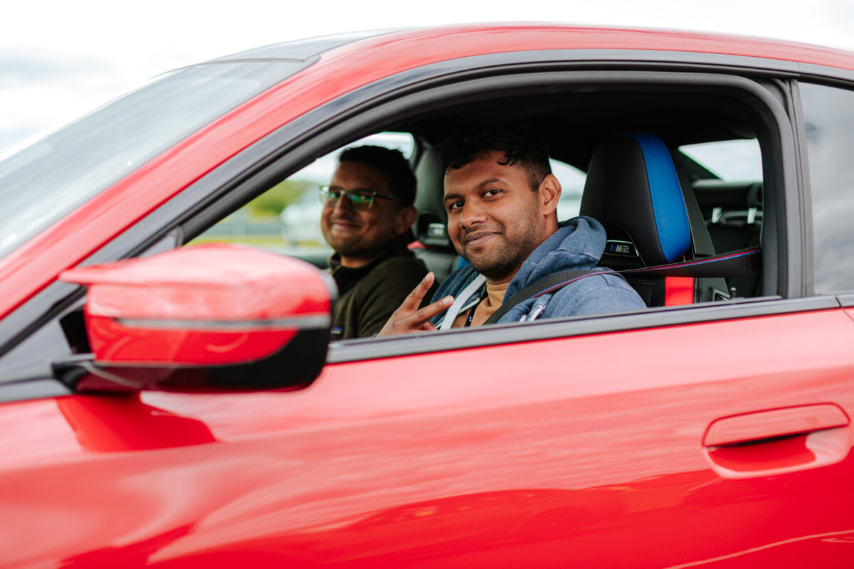 Two men are sitting in a Red BMW and one is displaying the Peace symbol.